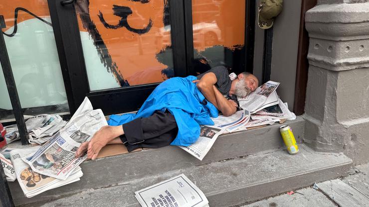 Man sleeps in a building entrance on newspapers in the morning in the commercial district of the Park Slope neighborhood of Brooklyn, New York on a summer morning.